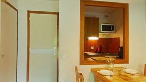 A dining area with a wooden table set for four, a kitchen visible through a doorway, and a white door. - Cozy studio - 4 people, parking, pets allowed (Le Grand-Bornand)