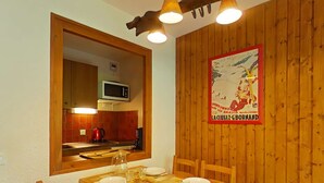 A dining area with a wooden table set for four, a wooden-framed mirror, and a kitchen visible in the background. - Cozy studio - 4 people, parking, pets allowed (Le Grand-Bornand)