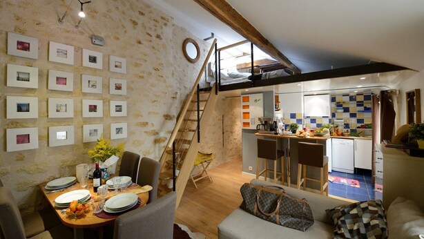 A dining area with a wooden table set for four, a staircase leading to an upper level, and a kitchen area visible through an open door.