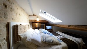 A cozy attic bedroom with a sloped ceiling, a skylight, a bed with pillows, a TV, and a bookshelf.