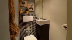 A bathroom with a white sink, a mirror, and a toilet. There are framed pictures on the wall and a small plant in a yellow pot.