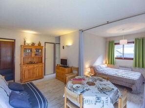 A compact room with a bed, a dining table set for two, a wooden cabinet, and a small table with a lamp.