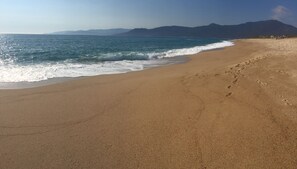 Une vaste plage de sable fin, avec des vagues douces, un ciel dégagé et des montagnes à l’horizon.