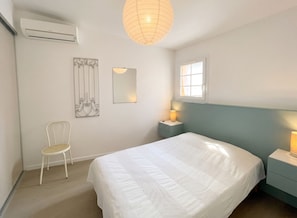 Un dormitorio con cama, una silla, una mesita de noche con lámpara, un espejo en la pared y un equipo de aire acondicionado.