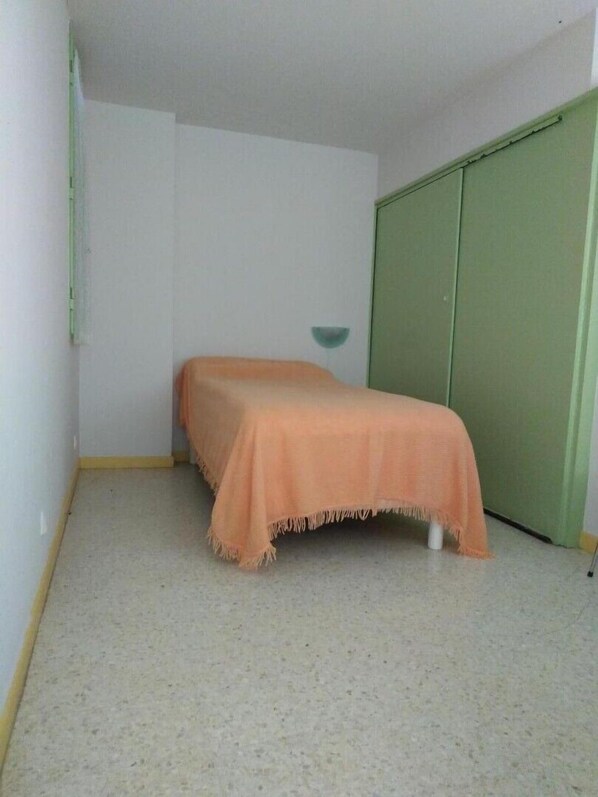 A small, sparsely furnished room with a single bed, a green wardrobe, and a light-colored floor.