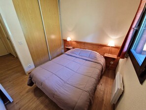 Quarto pequeno e bem organizado com uma cama de solteiro, cabeceira de madeira, mesas de cabeceira com abajures e uma janela com cortinas.