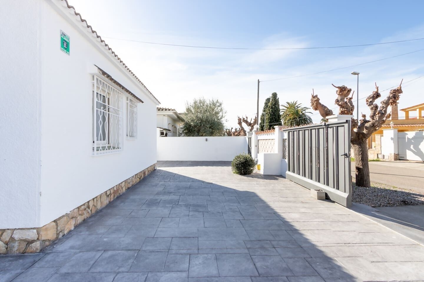 Un chemin pavé mène à une entrée fermée par un portail, avec un mur blanc et un portail en métal.