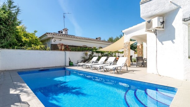 A swimming pool with tiled edges, surrounded by lounge chairs and a white building with stone pillars. - Piscina, mar y centro – Casa ideal para familias (Empuriabrava)