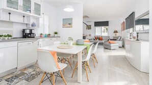 A modern kitchen with white cabinets, a dining area with a white table and chairs, and a living area with a sofa and a coffee machine. - Piscina, mar y centro – Casa ideal para familias (Empuriabrava)