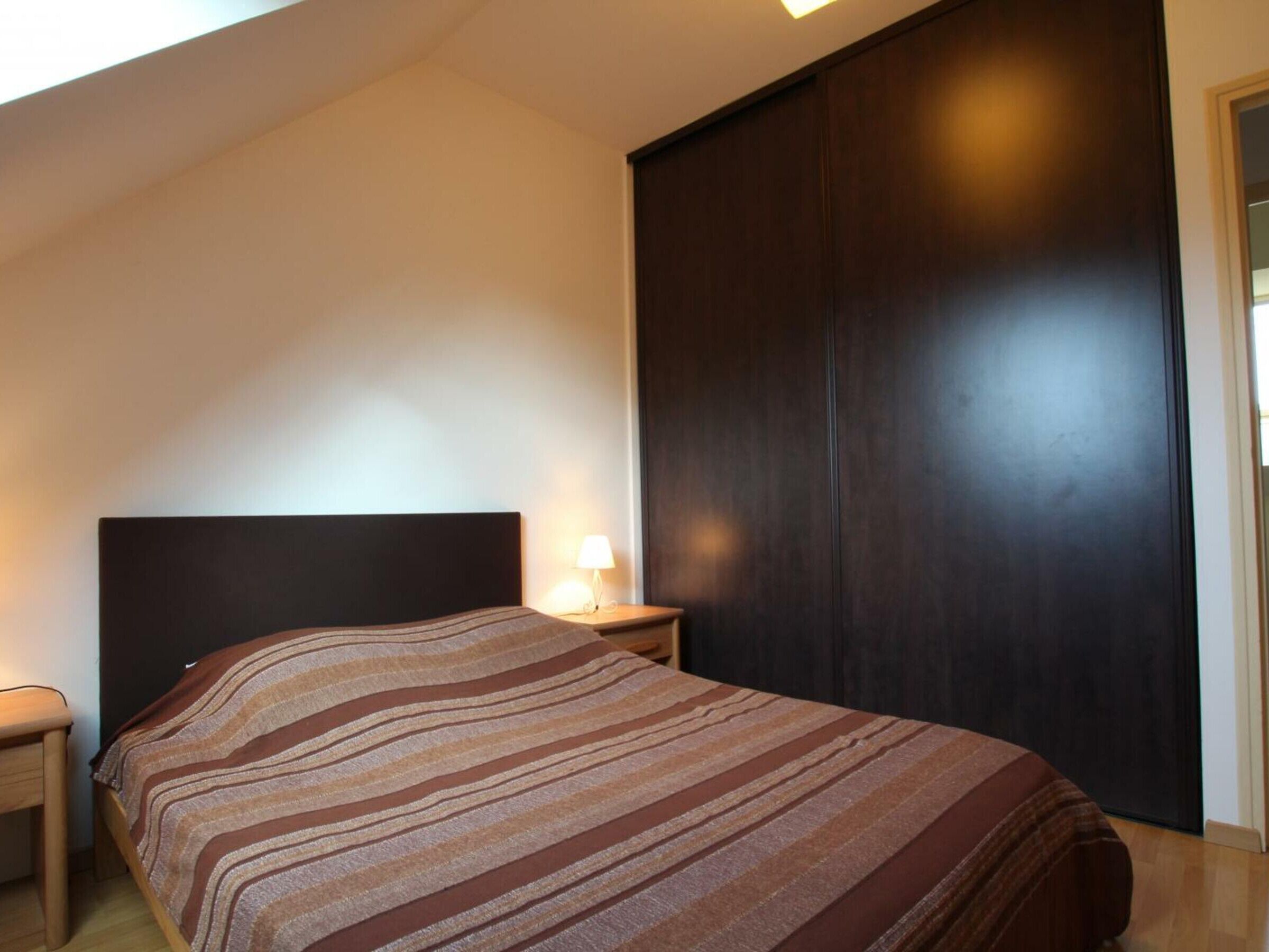 A bedroom with a bed, a nightstand, a lamp, and a dark wooden wardrobe.