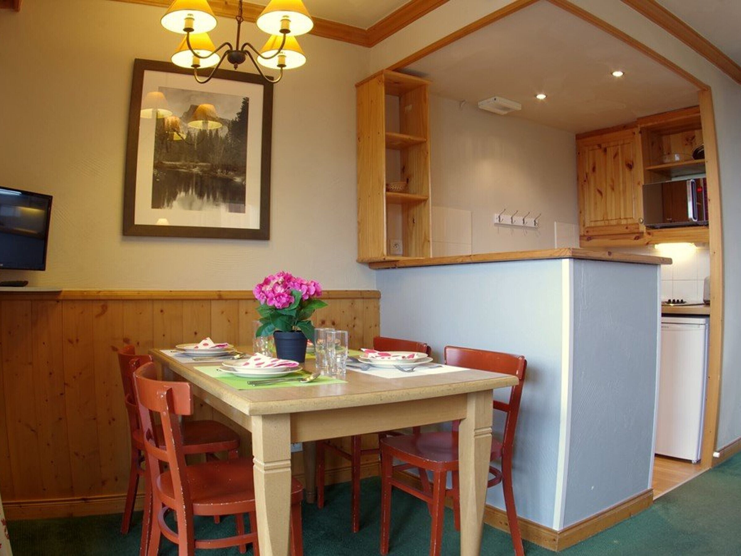 A dining area with a table set for four, wooden chairs, a vase with flowers, and a painting on the wall.