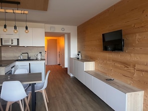 A modern kitchen with white cabinetry, a wooden wall, a built-in TV, and a dining area with a wooden table and chairs.
