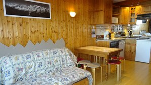 A cozy room with a wooden wall, a sofa with a patterned cover, a wooden table with two stools, and a kitchen area with wooden cabinets and appliances. - Studio 5 pers, ski-in/ski-out, near shops (Les Arcs 1800)