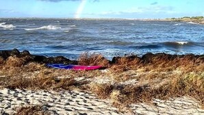 Uma praia com ondas, um arco-íris e um primeiro plano rochoso.