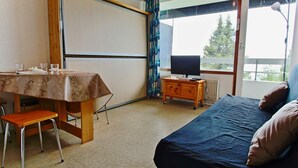 A room with a bed, a table set for two, a television on a wooden stand, and a window with a view of trees.