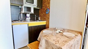 A compact kitchen with a microwave, coffee maker, and a small refrigerator. A dining table with a patterned tablecloth and cutlery is set for two.