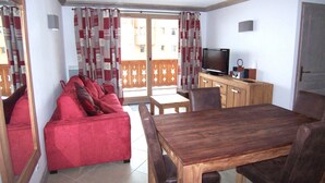 A room with a wooden dining table, a red sofa, a TV, and a balcony with patterned curtains.