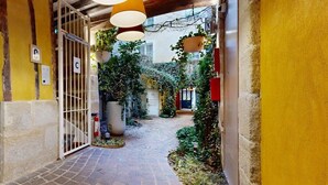A narrow alleyway with a brick floor, yellow walls, and a metal gate labeled 'C'. There are hanging plants and a red lamp above the gate.
