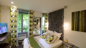A bedroom with a bed, a TV, a window with curtains, a wall with a leaf pattern, and a nightstand with a lamp.
