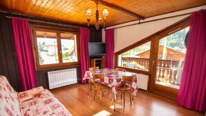 Una habitación acogedora con techo de madera, una mesa de comedor con sillas, un sofá, un televisor y ventanas grandes con cortinas.