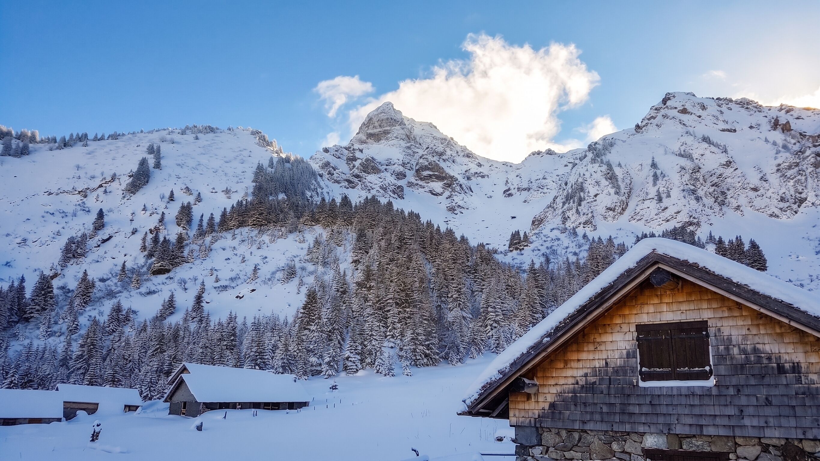 Een besneeuwd berglandschap met een houten chalet en een kleiner gebouw op de voorgrond.