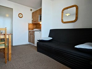 A compact living space with a kitchenette, a black sofa with pillows, a wooden chair, and a wall clock.