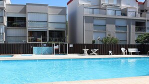 Une piscine avec une terrasse carrelée, entourée d’une clôture. On y trouve des chaises blanches et un banc près de la piscine.