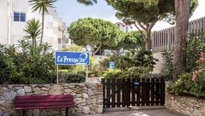 Une entrée fermée par un portail et bordée d’un mur de pierre, avec un banc et une pancarte indiquant « La Presqu’île ».