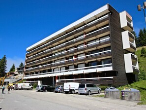 A multi-story building with balconies, parking area, and a small structure with a sign.