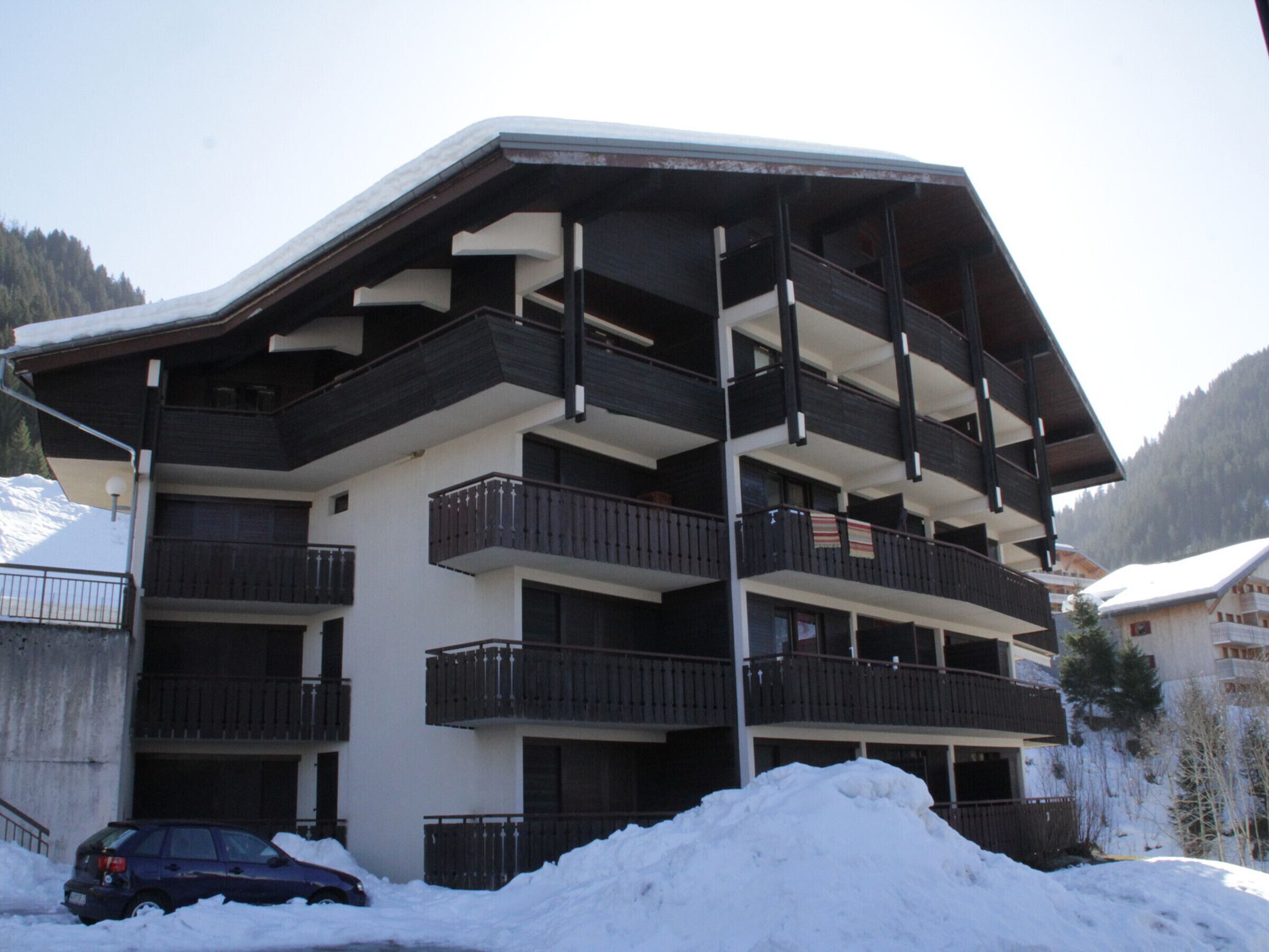 Un immeuble à plusieurs étages doté de balcons, une voiture garée devant et un sol recouvert de neige.