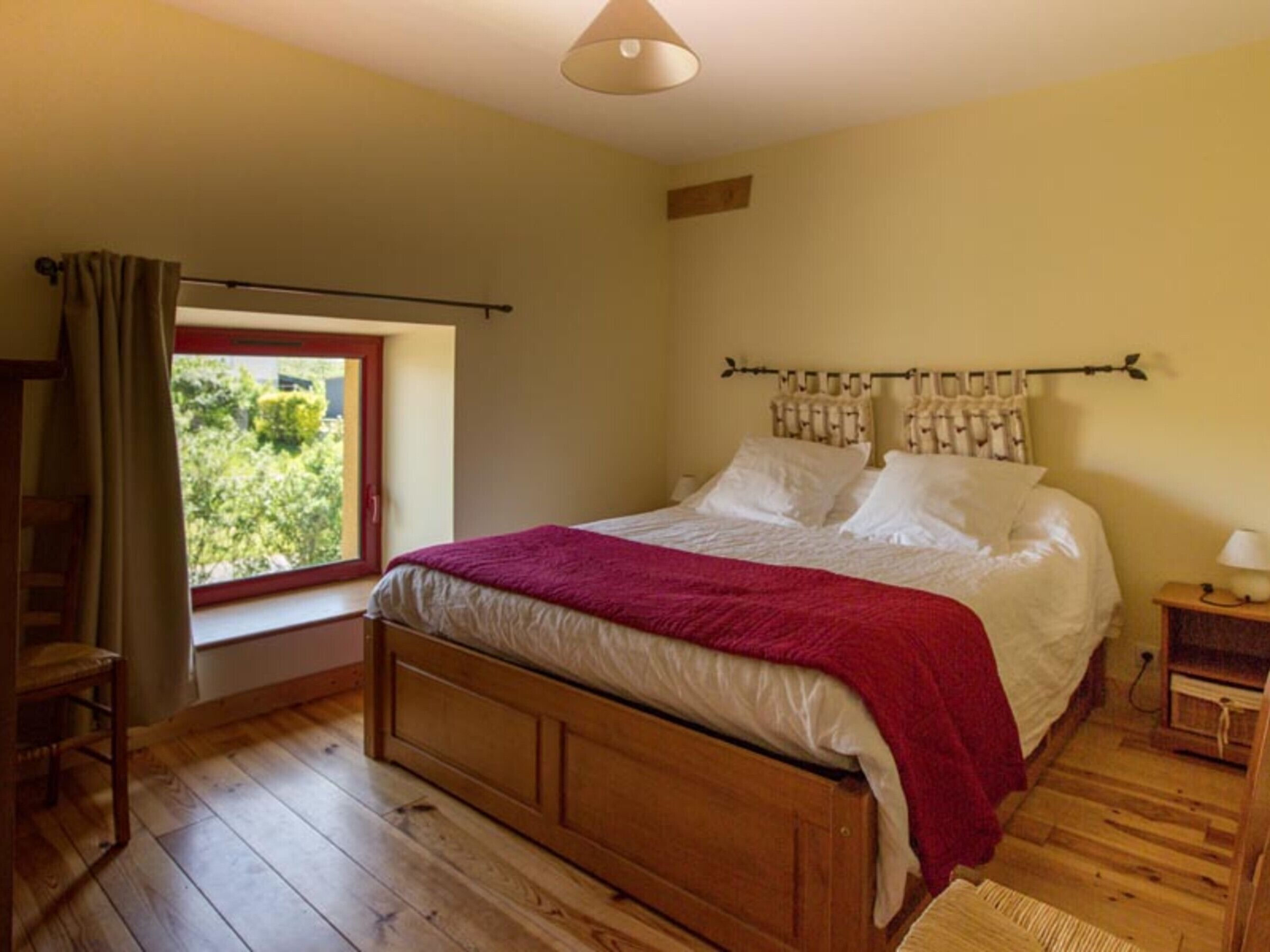 A bedroom with a wooden bed, a window with a view of greenery, a wooden nightstand with a lamp, and a chair.