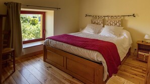 A bedroom with a wooden bed, a window with a view of greenery, a wooden nightstand with a lamp, and a chair. - Large house with heated pool and spa in Manche (Savigny (Manche))
