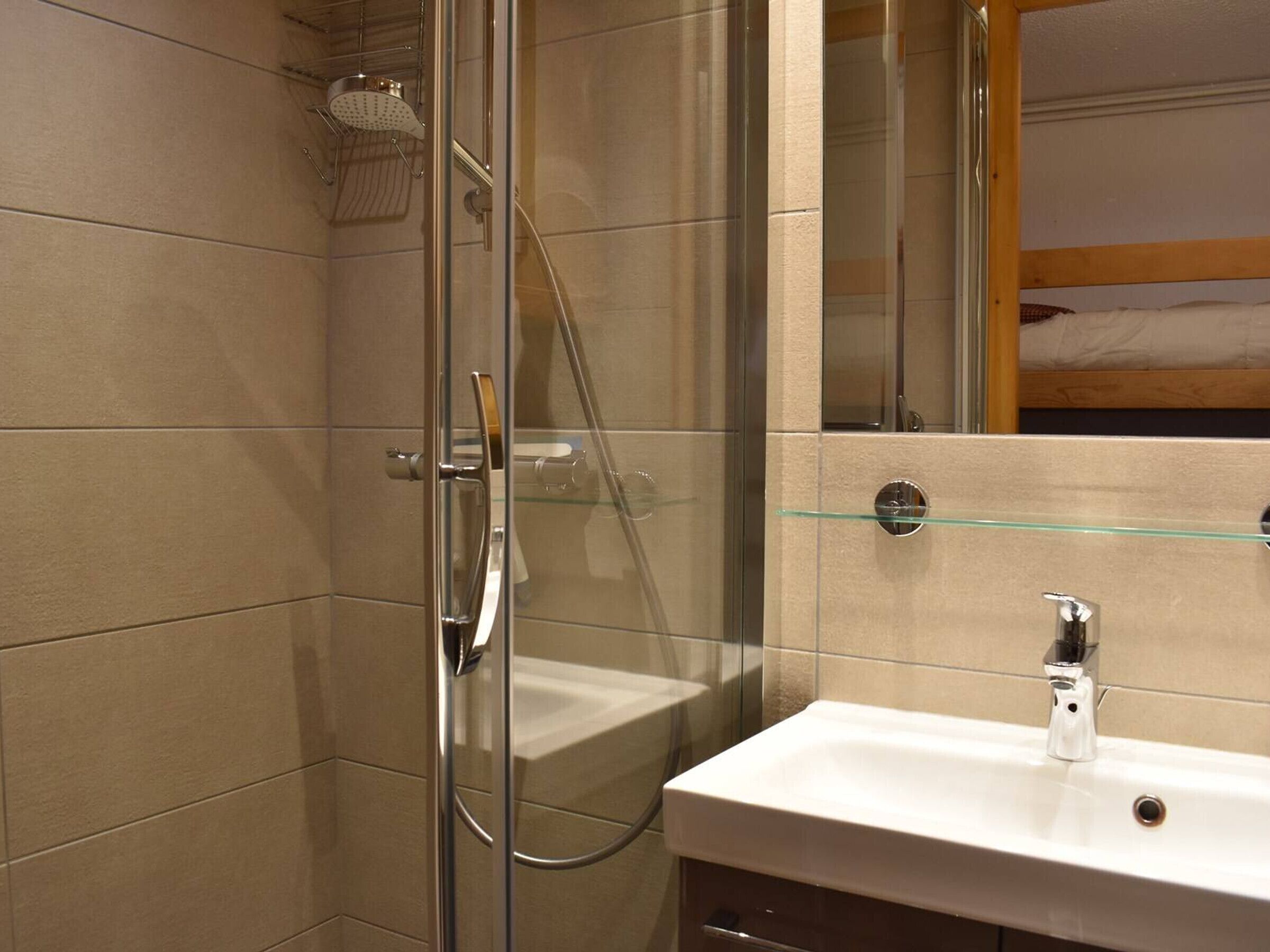 Ein modernes Badezimmer mit Duschkabine aus Glas, einer Glasablage über dem Waschbecken und einem Holzbett im Hintergrund.