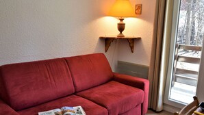A red sofa with a magazine, a table with a yellow lamp, and a window with a view of trees. - Calm mountain corner studio, 4 people, parking, Risoul 1850 (Risoul)