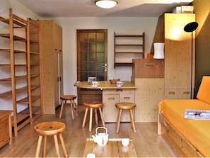 Quarto compacto com uma cama, pequeno espaço de cozinha, guarda-roupa de madeira e um banquinho de madeira.
