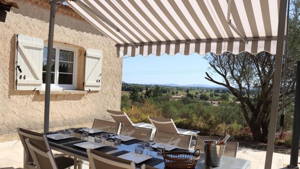 An outdoor dining area with a table set for four, surrounded by chairs, under a striped awning, overlooking a scenic landscape.