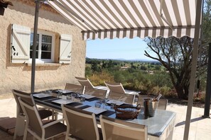 Un área de comedor al aire libre con una mesa dispuesta para cuatro personas, rodeada de sillas, bajo un toldo a rayas, con vistas a un paisaje pintoresco.