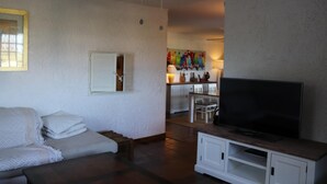 A living room with a sofa, a TV on a wooden stand, a ceiling fan, and a kitchen area in the background.