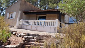A two-story house with a balcony, surrounded by natural vegetation and a stone pathway leading to the entrance.