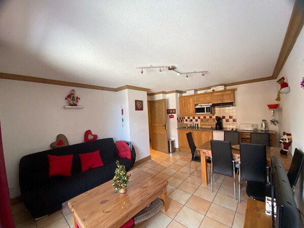 A cozy living room with a black sofa, red cushions, and a small Christmas tree on a wooden coffee table. A kitchen with wooden cabinets and a microwave is visible in the background.