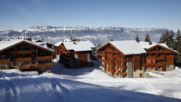 Une rangée de cabanes en bois aux toits enneigés, se dressant sur fond de montagnes aux sommets couverts de neige et d’un ciel dégagé.