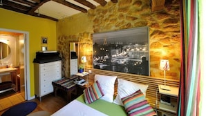 A bedroom with a stone wall, a bed with colorful pillows, a nightstand with a lamp, and a large framed picture of a cityscape. - B3DD Honeymoon (París)
