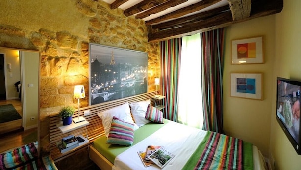 A bedroom with a stone wall, wooden beams, a bed with colorful bedding, a nightstand, a lamp, and a framed picture on the wall. - B3DD Honeymoon (París)