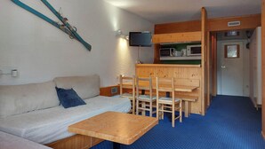 Un espacio habitable compacto con un sofá, una pequeña mesa de madera, un televisor montado en la pared y una kitchenette con microondas y cafetera.