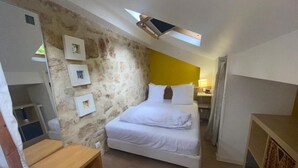 A bedroom with a stone wall, a bed with white bedding, a wooden nightstand, and a skylight above the bed.
