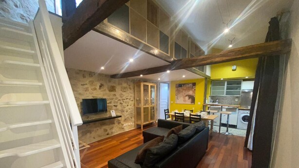 A modern living room with a black sofa, wooden flooring, a stone wall, and a kitchen area with a yellow accent wall.