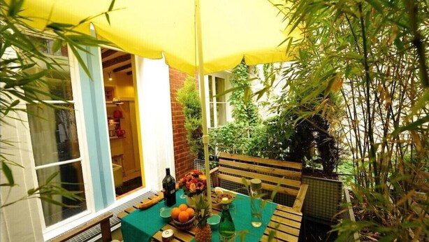 A patio with a table set for two, a yellow umbrella, and a view of a brick wall and greenery. - E1GG Everglades (Paris)