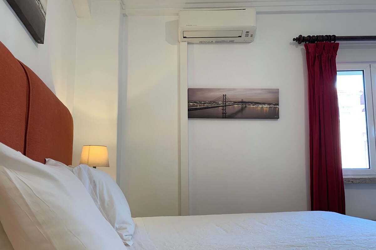 A hotel room with a bed, a wall-mounted air conditioner, a painting of a bridge, and a window with red curtains.