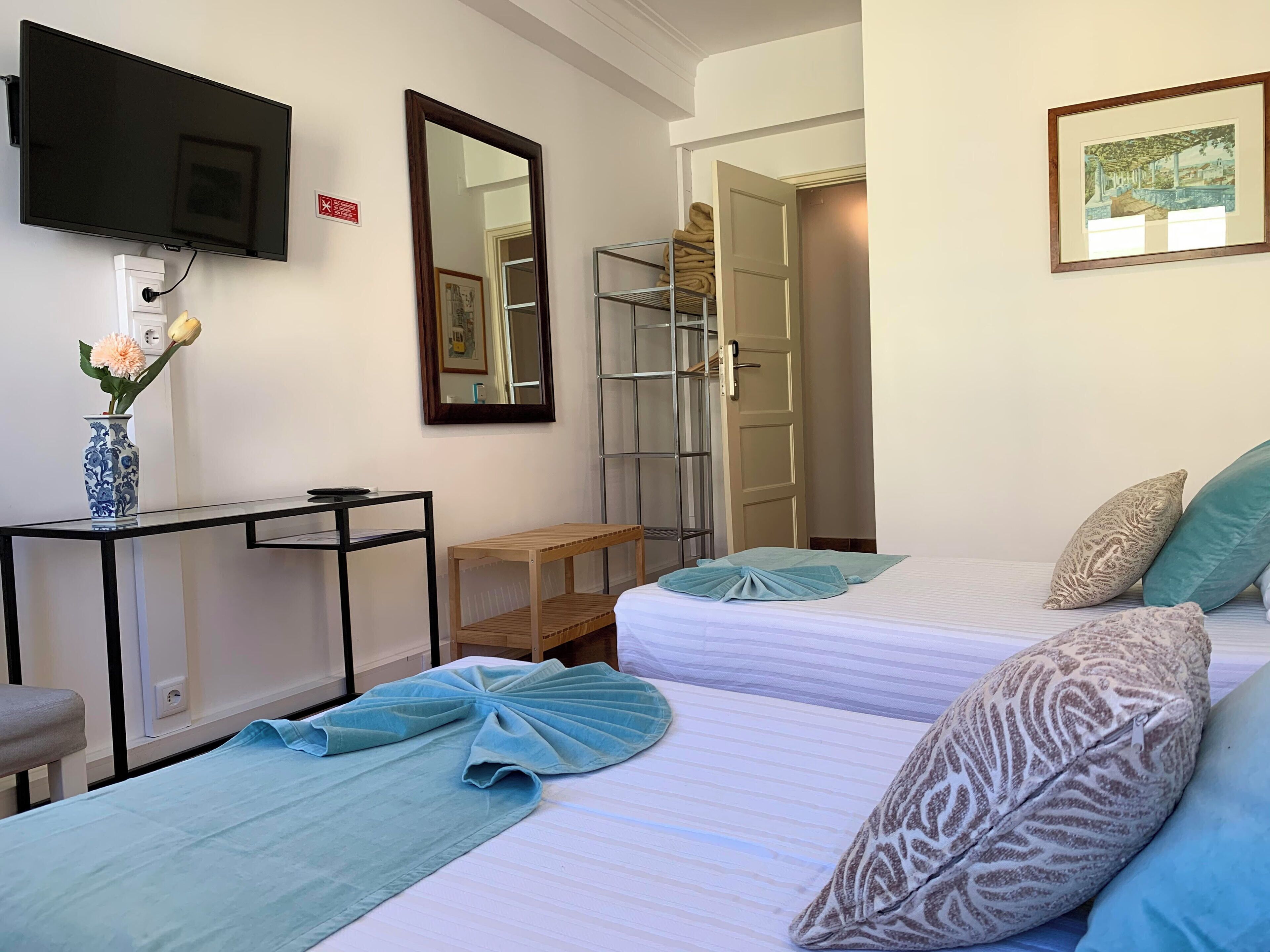 Ein Hotelzimmer mit zwei Betten, einem Fernseher, einem Spiegel, einem kleinen Tisch mit einer Vase und einem Kleiderschrank.