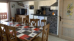 A compact kitchen with white cabinets, a microwave, and a sink. There is a dining area with a table and chairs, and a TV on a stand.